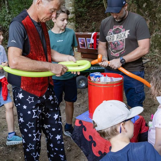 Ein Clown macht Luftballontiere für Kinder in einer Waldumgebung, mit einem Mann in einem grauen T-Shirt und einem roten Eimer in der Nähe.