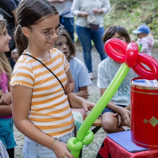 Ein junges Mädchen hält eine Ballonfigur und lächelt, während sie sie ansieht. Hinter ihr beobachten andere Kinder und Erwachsene. Der Ort scheint im Freien zu sein.