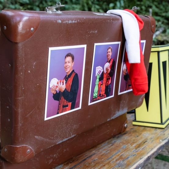 Ein brauner Koffer mit drei Fotos eines Zauberers darauf. Der Zauberer hält ein Kartenspiel und ein roter Schal liegt auf dem Koffer.