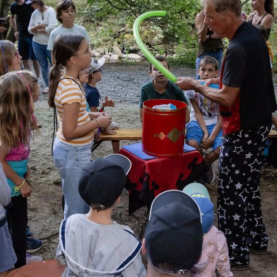 Ein Zauberer führt einen Ballontrick für eine Gruppe von Kindern im Freien vor. Die Kinder beobachten aufmerksam, und einige Erwachsene stehen in der Nähe.