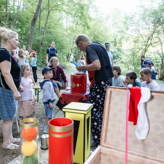 Ein Zauberer führt einen Trick mit einem roten Eimer und einem Hut in einem Wald vor, umgeben von einer Menge Kinder und Erwachsener.
