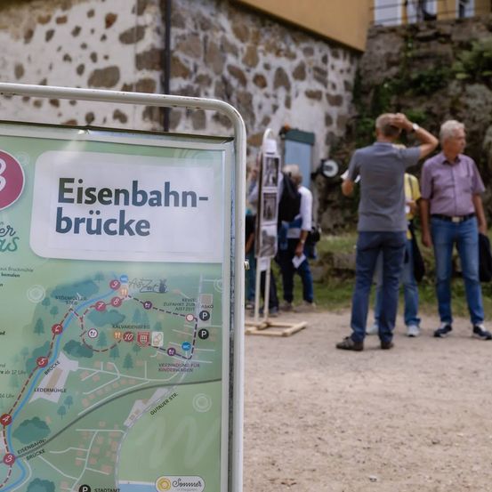 Ein Kartenschild für die Eisenbahn-brucke mit einigen Personen im Hintergrund, von denen eine ein Foto macht.