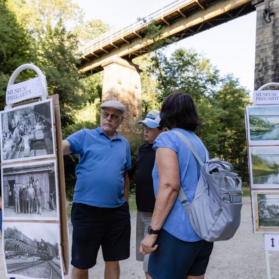 Ein Mann und eine Frau stehen vor einem Fotodisplay, mit einer Brücke und Bäumen im Hintergrund.