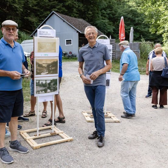 Eine Gruppe älterer Männer und Frauen steht um eine Holztafel mit alten Fotos herum, lächelt und unterhält sich in einem Park.