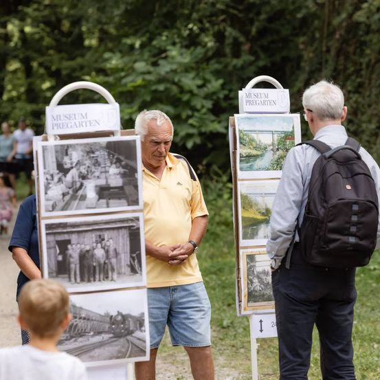 Ein älterer Mann in einem gelben T-Shirt steht vor einer Fotoausstellung, während ein Mann mit Rucksack zusieht. Die Ausstellung hat mehrere Schwarz-Weiß-Fotos und Schilder, die Museum Pregarten lesen.