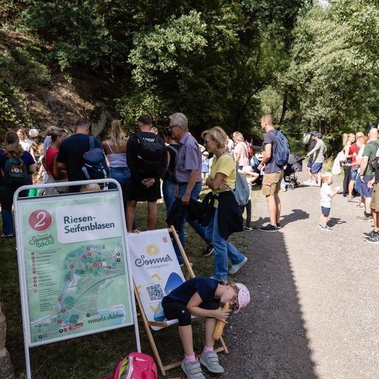 Eine Menschenmenge, einige mit Rucksäcken, versammelt sich in einem Park, mit einem Kartenschild und einem Kind auf einem Stuhl sitzend.