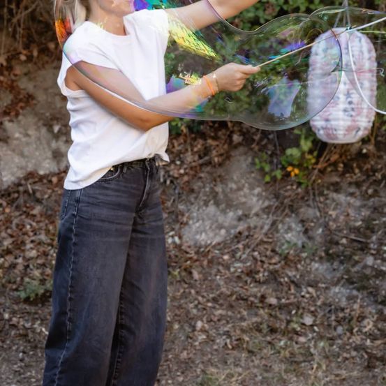 Eine Frau in einem weißen T-Shirt und blauen Jeans bläst draußen eine große, bunte Blase.