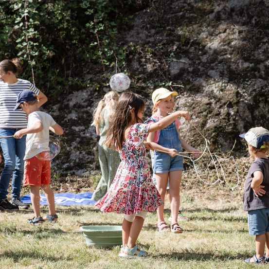 Eine Gruppe von Kindern spielt im Freien, einige mit Seifenblasenstäben, in einem Grasbereich mit einer Steinmauer im Hintergrund.