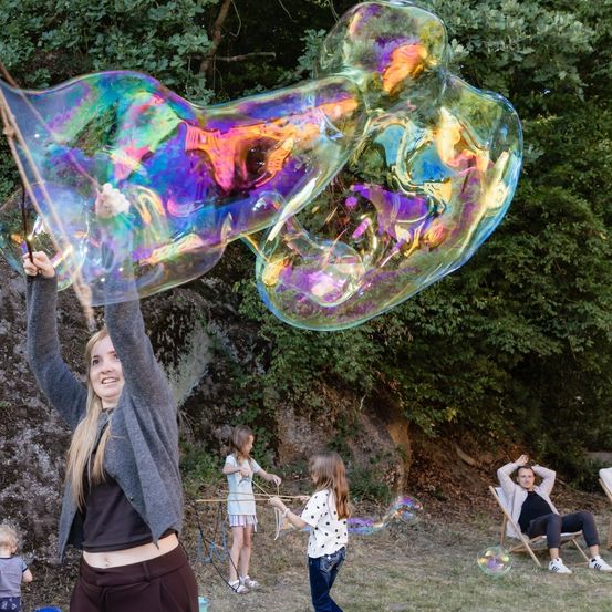 Eine Frau hält eine große, bunte Seifenblasen-Stange und bläst eine riesige Seifenblase in einem Park, während Kinder und Erwachsene in der Nähe Seifenblasen genießen.
