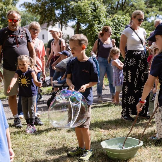 Eine Gruppe von Menschen, einschließlich Kindern, versammelt sich in einem Park. Ein Junge hält eine große Seifenblasen-Wand und ein Mädchen neben ihm beobachtet, wie er eine Blase bläst.