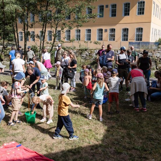 Eine Gruppe von Menschen, einschließlich Kindern, versammelt sich auf einer Rasenfläche vor einem Gebäude. Einige beugen sich nach unten, möglicherweise bei einer Aktivität.
