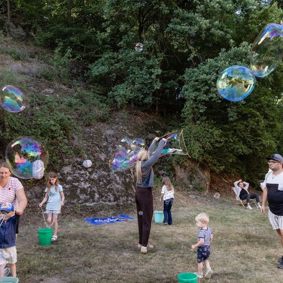 Eine Gruppe von Menschen bläst Seifenblasen in einem Grasfeld mit Bäumen im Hintergrund. Einige halten Blasenringe, während andere die Blasen schweben sehen.