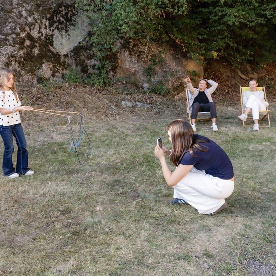 Drei Personen sitzen in Stühlen, während eine mit einem Drachen spielt. Eine weitere Person kniet und macht ein Foto.