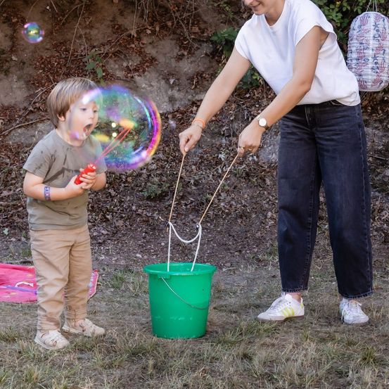 Eine Frau und ein kleiner Junge spielen mit einem Seifenblasenstab im Garten. Der Junge bläst Seifenblasen und die Frau hält einen Eimer.