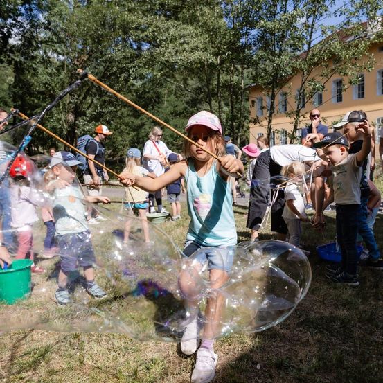 Eine Gruppe von Kindern spielt mit Seifenblasenstäben auf einer Wiese, mit einem Gebäude und Bäumen im Hintergrund.