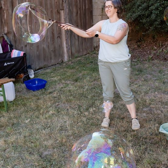 Eine Frau im Garten bläst mit einem Stock eine große Seifenblase. Sie lächelt und trägt ein ärmelloses Oberteil und Hose.