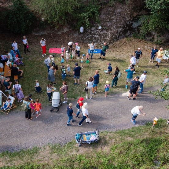 Eine Luftaufnahme einer großen Gruppe von Menschen, die sich in einem Park versammelt haben, einige stehen, andere sitzen auf Bänken, während Kinder auf dem Gras spielen.