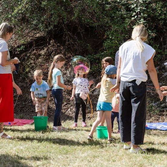 Eine Gruppe von Kindern und Erwachsenen spielt im Freien. Die Kinder halten grüne Eimer und eine Seifenblasen-Stange, während eine erwachsene Frau eine Seifenblasen-Stange hält. Sie stehen auf einem Grasfeld mit einer Steinmauer im Hintergrund.