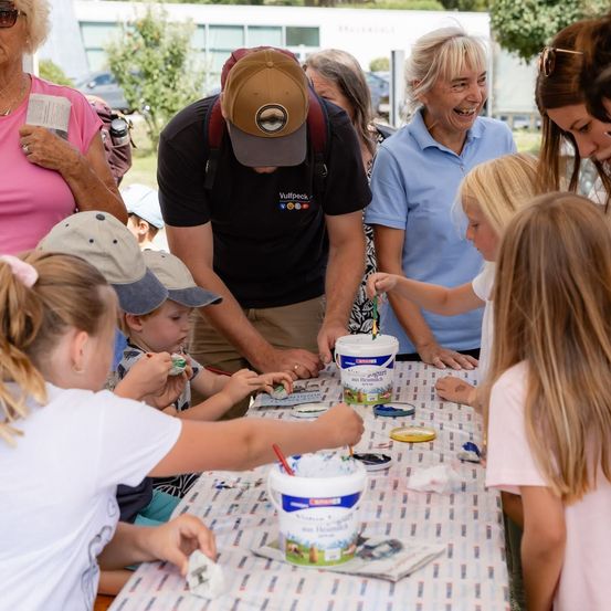 Eine Gruppe von Kindern und Erwachsenen versammelt sich um einen Tisch und ist in einer Aktivität vertieft. Die Kinder malen oder verzieren etwas auf dem Tisch, während Erwachsene zusehen und helfen.