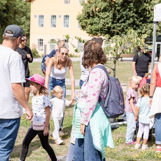 Eine Gruppe von Menschen, einschließlich Erwachsener und Kindern, geht auf einem Rasen mit einem Gebäude im Hintergrund. Einige tragen Hüte, Brillen und Rucksäcke.