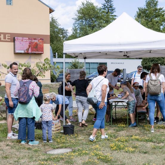 Eine Gruppe von Menschen, einschließlich Kinder, versammelt sich um einen Tisch unter einem Zelt in einem Park, mit einem Gebäude, das ein digitales Schild im Hintergrund anzeigt.