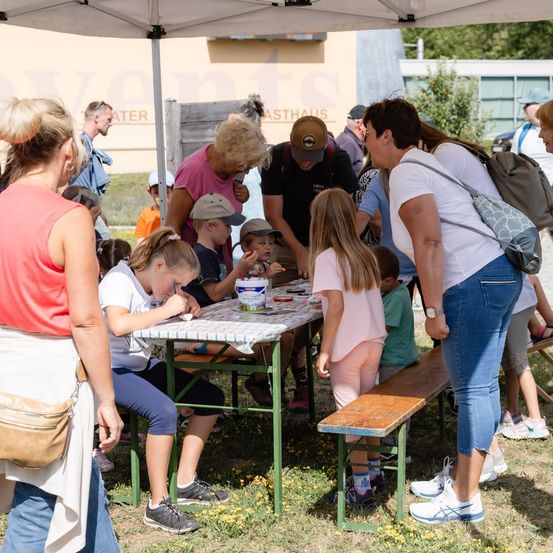 Eine Gruppe von Erwachsenen und Kindern versammelt sich um einen Tisch unter einem Zelt, wobei einige an Aktivitäten beteiligt sind und andere zusehen. Auf dem Tisch stehen ein Eimer und verschiedene Gegenstände.