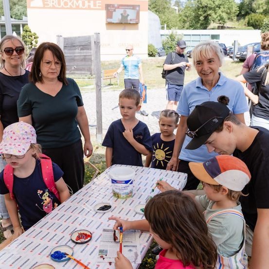Eine Gruppe von Erwachsenen und Kindern versammelt sich um einen Tisch und malt mit Joghurtbechern und Pinseln. Die Erwachsenen lächeln und beobachten die Kinder. Im Hintergrund steht ein Gebäude.