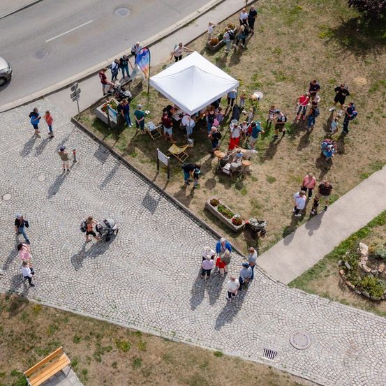 Eine Luftaufnahme einer Gemeinschaftsveranstaltung mit einem weißen Zelt, Menschen, die gehen, und einem Kinderwagen. Die Umgebung umfasst einen gepflasterten Bereich, Grasflächen und eine Straße.