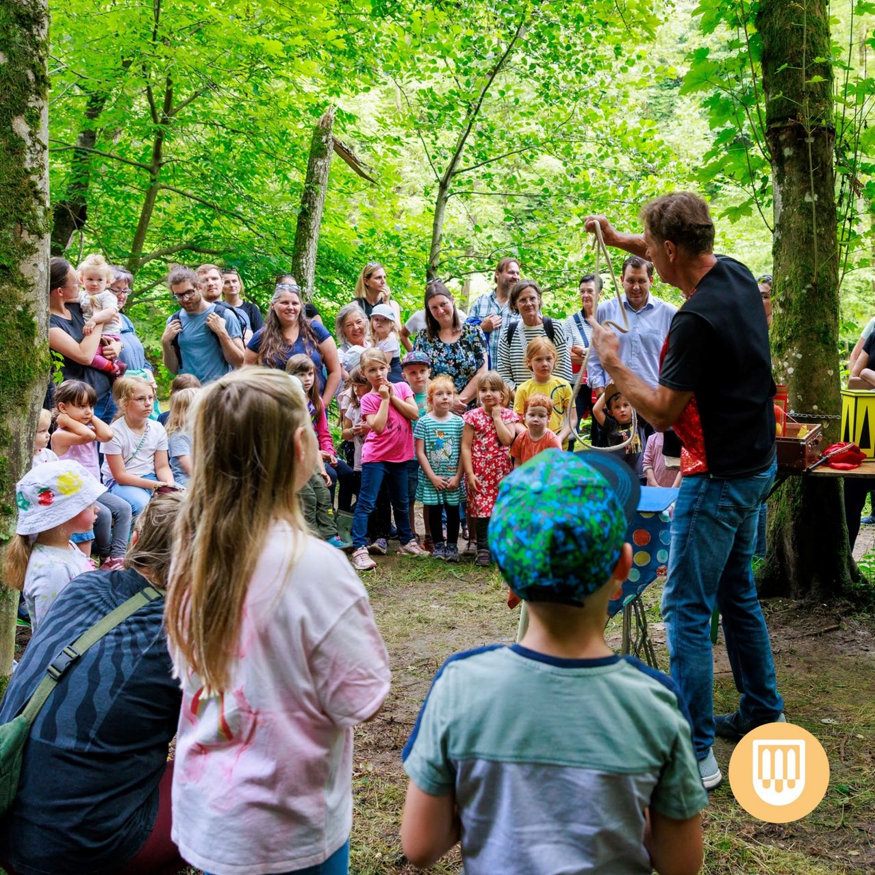Eine Gruppe von Erwachsenen und Kindern versammelt sich im Wald. Ein Mann führt mit einem Seil vor, und ein kleines Publikum schaut zu.