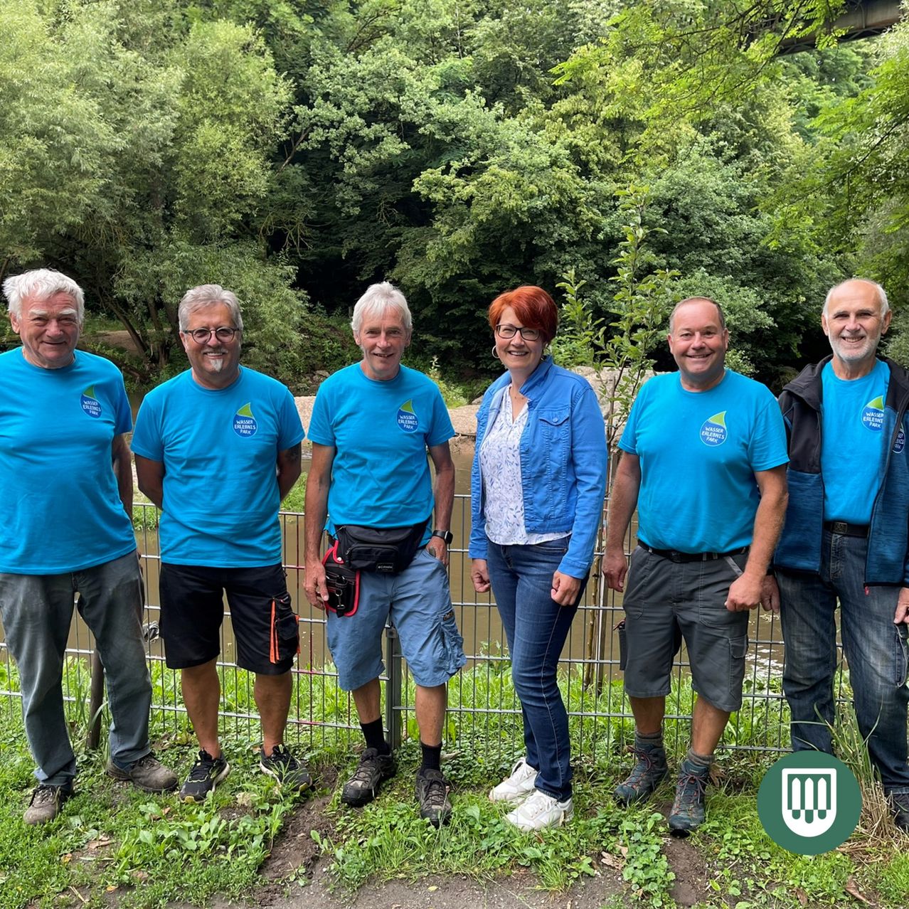 Sechs Menschen stehen zusammen vor einem Zaun und lächeln für ein Foto. Sie tragen alle blaue Shirts, und einige haben zusätzliche Accessoires. Dahinter befindet sich eine natürliche Umgebung mit Bäumen und Gras.