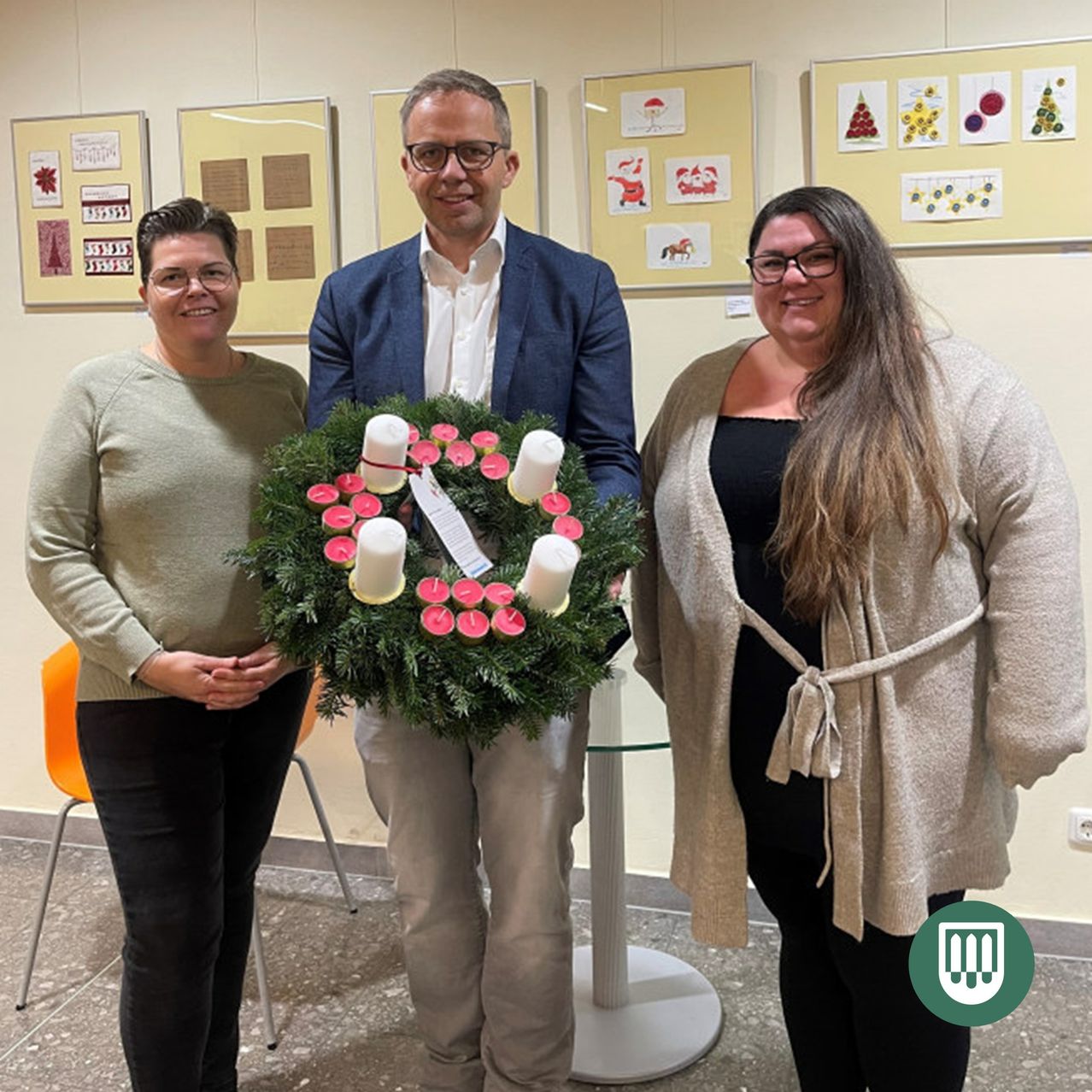 Drei Menschen stehen in einem Raum, einer Mann hält eine Kerzenkranz. Die Frauen auf beiden Seiten lächeln in die Kamera. Hinter ihnen hängen verschiedene gerahmte Bilder an der Wand.