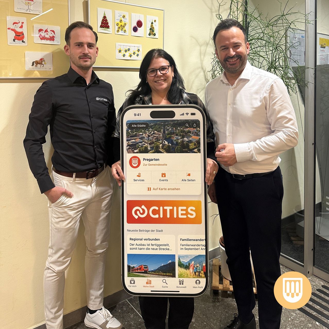 Drei Personen posieren mit einem Telefon in einem Konferenzraum. Das Telefon zeigt die App 'cities'. Die Leute lächeln und scheinen zu feiern.