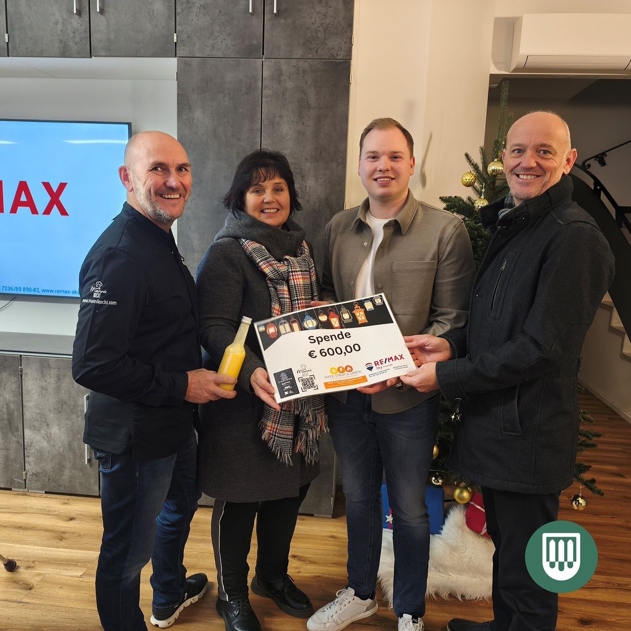 Vier Personen stehen in einem Raum, lächeln und haben einen Weihnachtsbaum hinter sich. Ein Mann hält eine Geschenkbox mit einem Zettel von 600,00 € und einer Flasche Saft. Der Raum hat Holzböden und graue Wände. Ein Monitor mit dem Wort 'MAX' ist sichtbar.