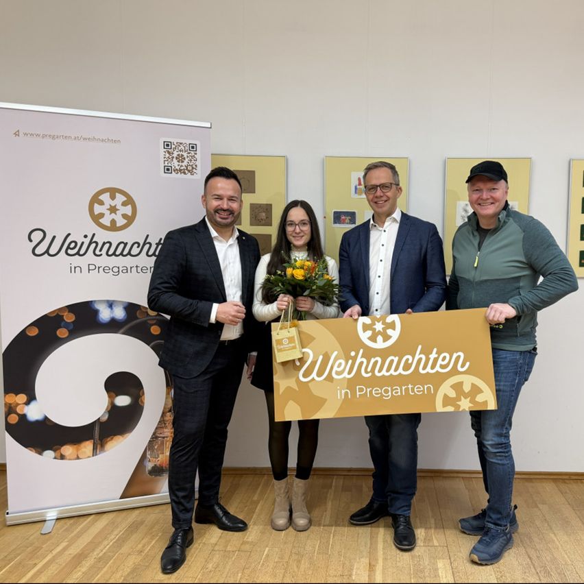 Vier Personen stehen in einem Raum zusammen. Sie halten ein großes Schild mit der Aufschrift 'Weihnachten in Pregarten'. Die Frau in der Mitte hält einen Blumenstrauß und einen Beutel. Der Raum hat einen Holzboden und eine weiße Wand.