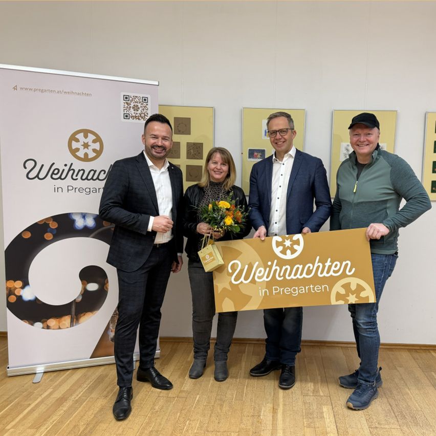 Vier Personen stehen lächelnd in einem Raum und halten ein Schild mit 'Weihnachten in Pregarten' darauf. Die Frau in der Mitte hält einen Blumenstrauß. Hinter ihnen sind Banner ausgestellt, und der Raum hat Holzboden.