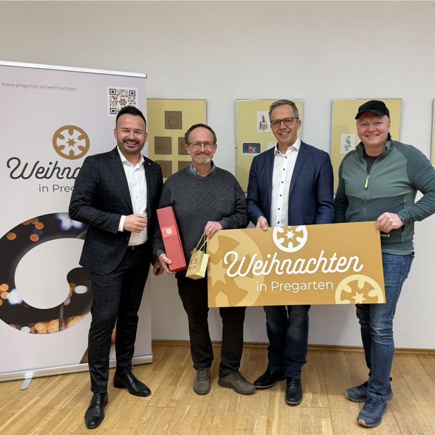 Vier Männer stehen vor einem Schild mit der Aufschrift 'Weihnachten in Pregarten'. Sie halten eine Geschenkbox und eine Plakette und lächeln für ein Foto. Dahinter befindet sich ein Banner mit demselben Text.