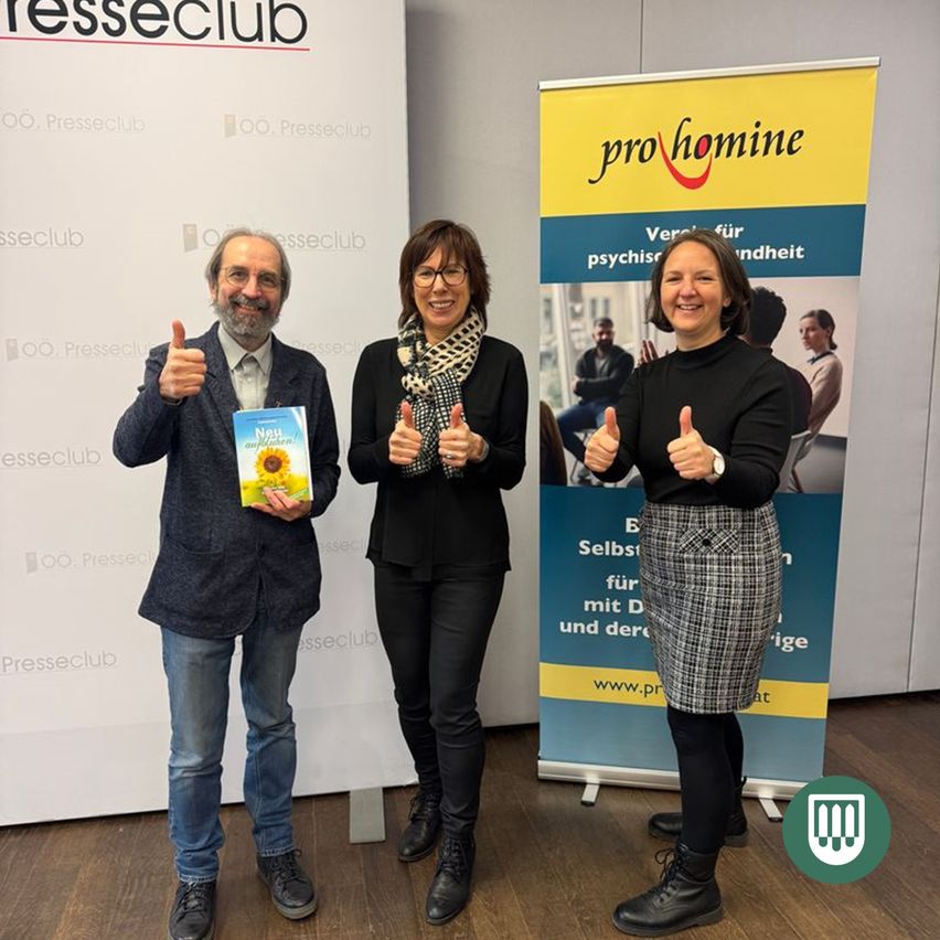 Drei Personen stehen vor einem Pressclub-Banner, lächeln und zeigen Daumen hoch. Der Mann hält ein Buch mit dem Titel Neu und Alter. Dahinter befindet sich ein pro HOMINE-Banner.