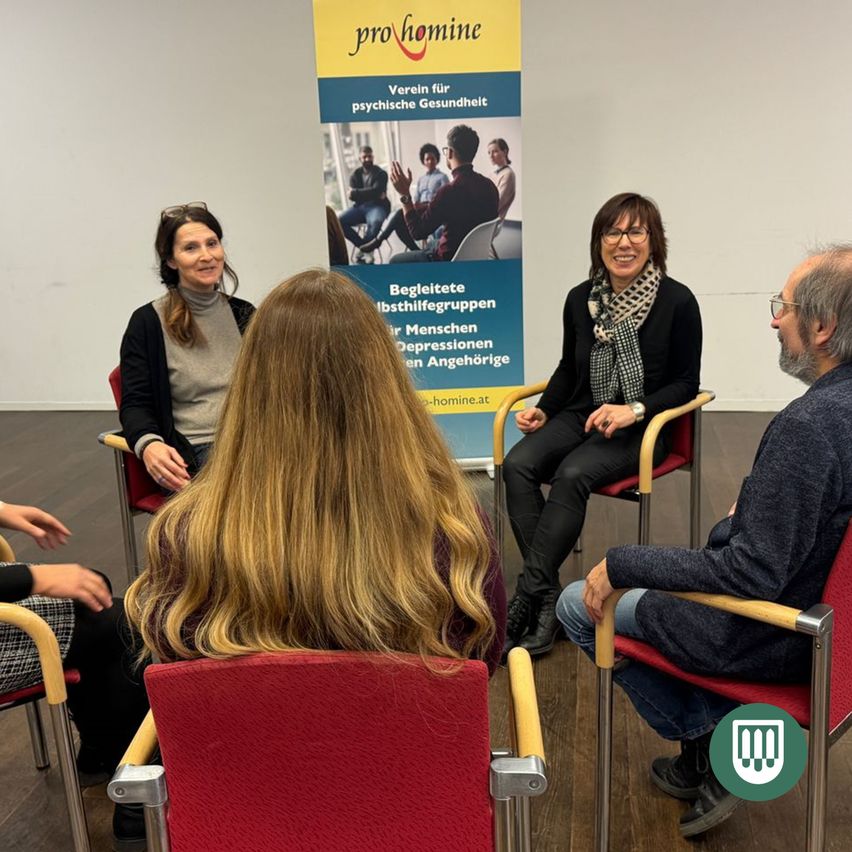 Fünf Personen sitzen in Stühlen, möglicherweise in einer Besprechung. Sie lächeln und schauen sich gegenseitig an. Hinter ihnen befindet sich ein Banner mit einem Logo und Text.