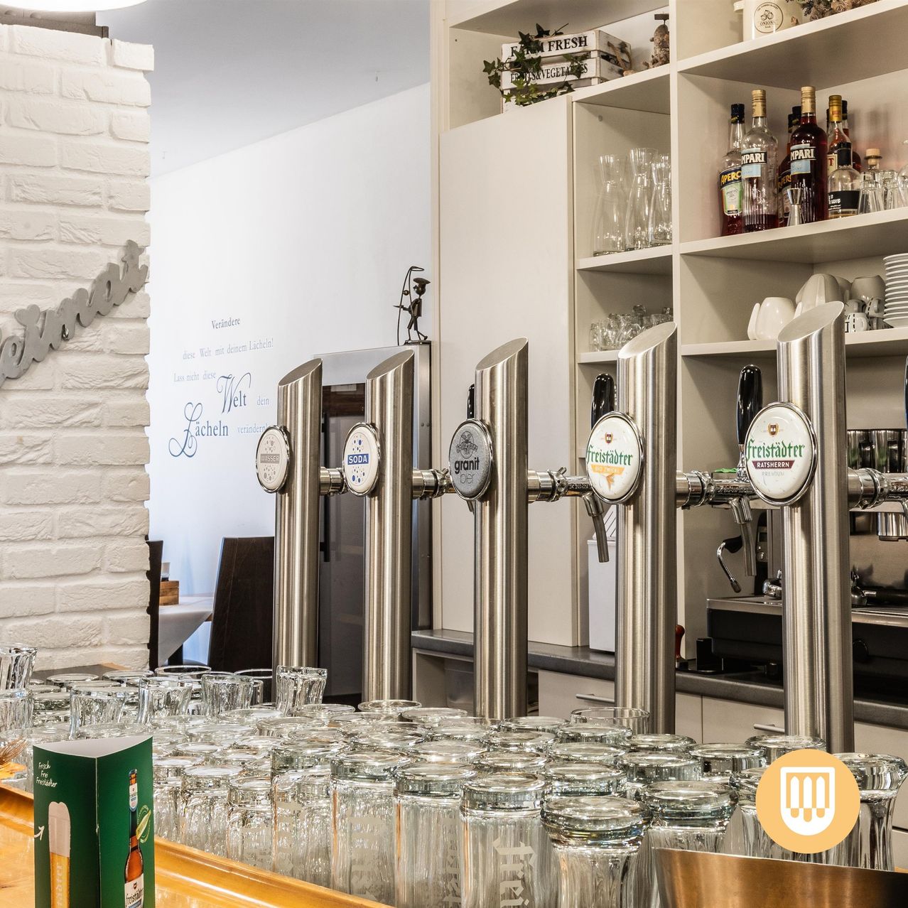 The interior of a bar or restaurant with stainless steel beer taps labeled with various brands. The background features a white brick wall and shelves stocked with bottles and glasses. The foreground shows a table with numerous glasses and a green box.