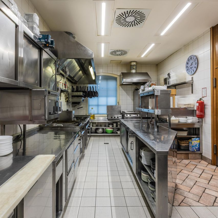 A commercial kitchen with stainless steel appliances, white tiled flooring, and a fire extinguisher. The kitchen has a window with a blue frame and a clock on the wall.