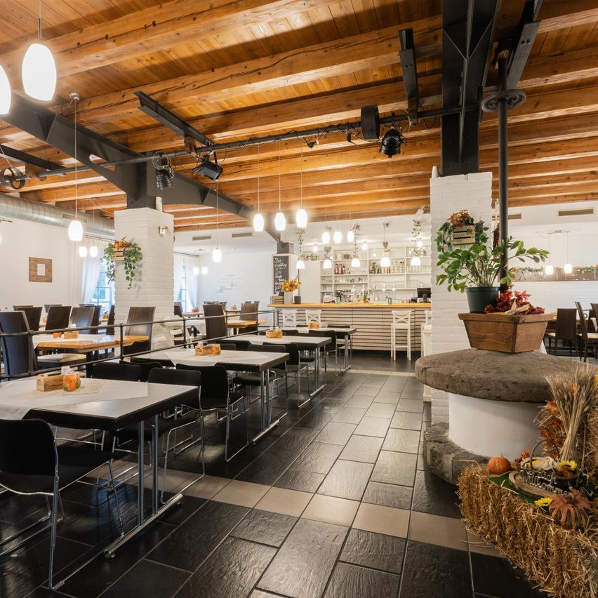 A modern restaurant with wooden beams, tables, and chairs. A counter with plants and autumn decorations. A chandelier and spotlights. A wooden barrel with a plant.