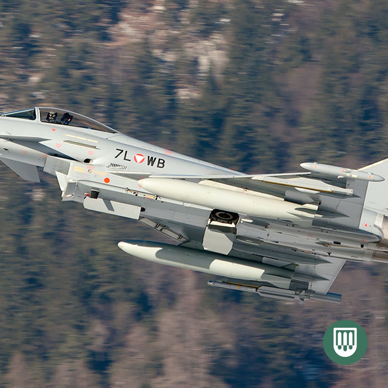 A military jet with the number 7L WB is flying over a mountainous region with snowy peaks.