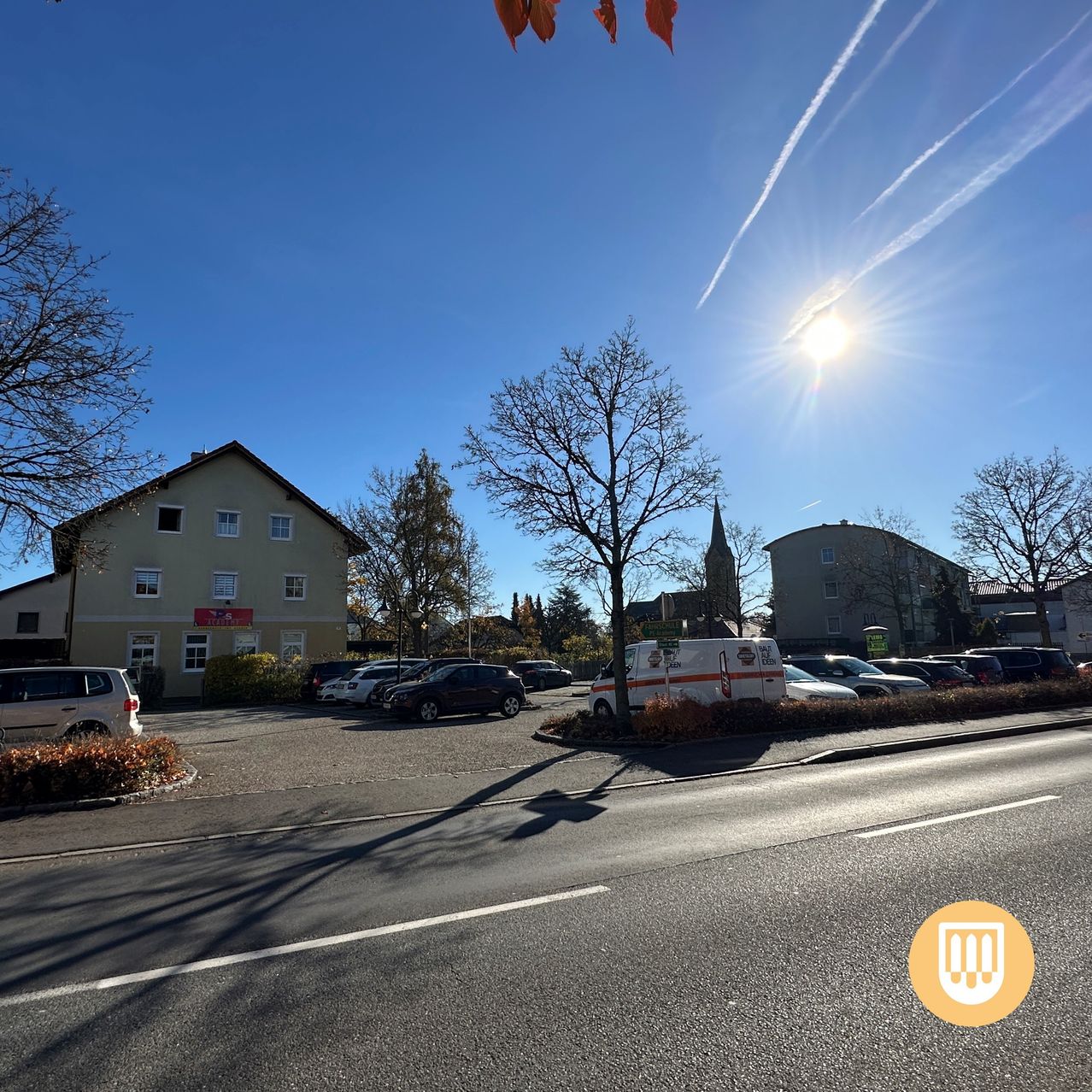 A sunny day shows a parking lot with several cars parked. The building has a red sign, and a tree is beside it. In the distance, another building is visible.