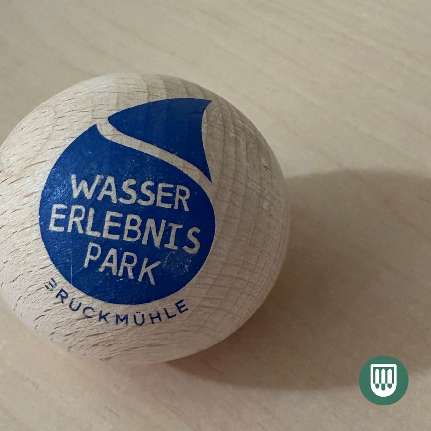 Ein Holzball mit dem Text 'Wassererlebnis Park' darauf. Es hat einen blauen Kreis mit einem Wassertropfen-Design. 'Ruckmuhle' steht in weißer Schrift unten.
