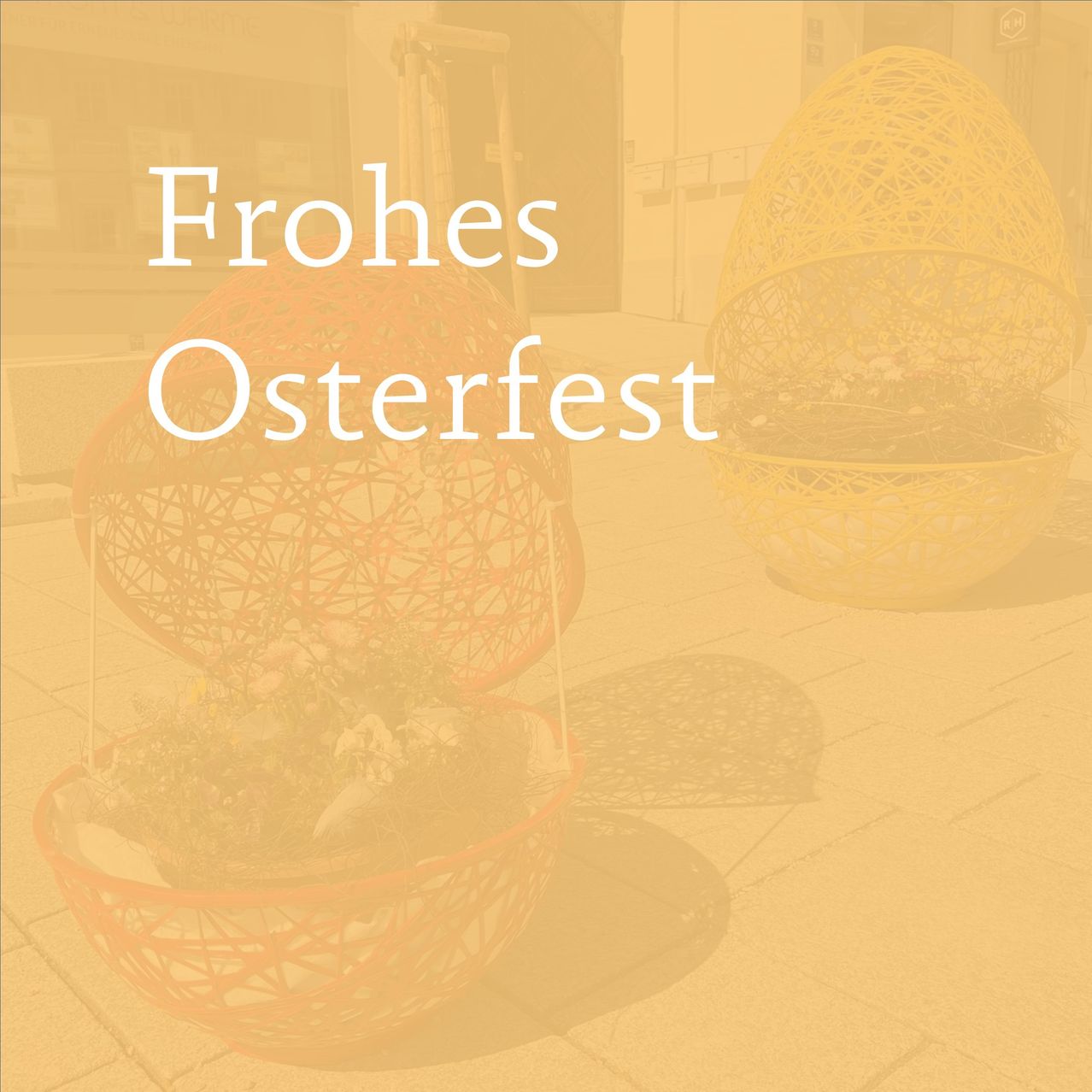 Zwei dekorative Eier auf einem Gehweg. Eines ist offen mit Blumen im Inneren. Im Hintergrund stehen die Worte Frohes Osterfest.