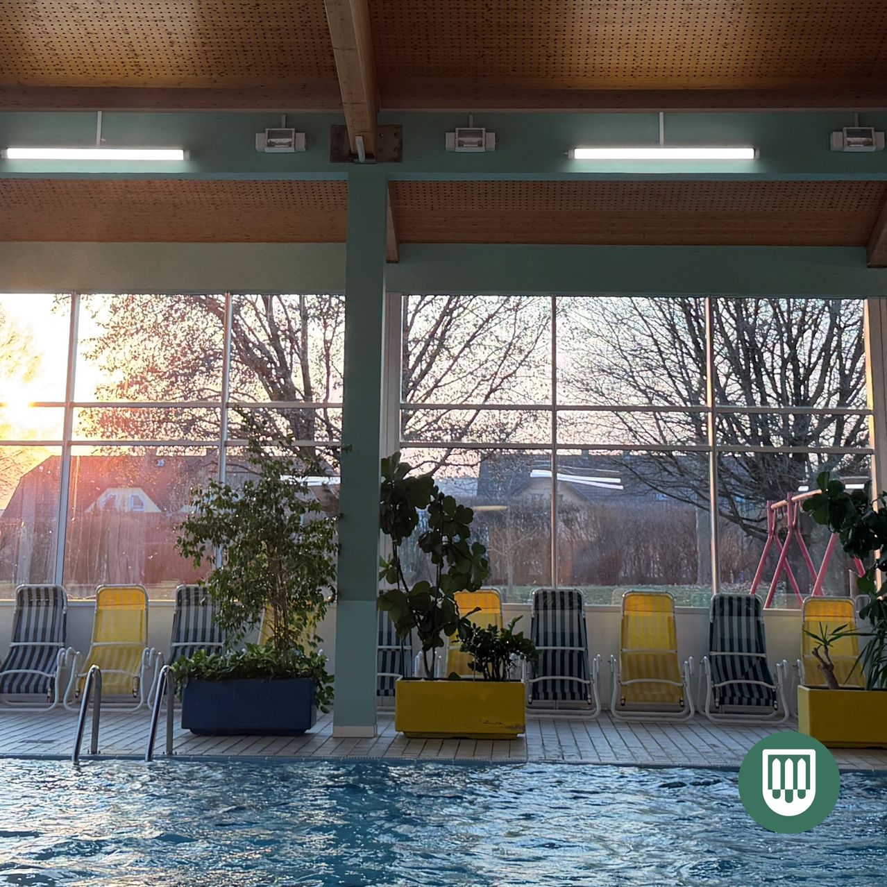 Ein Hallenbad mit sonnendurchflutetem Ausblick durch große Fenster. Gelbe und blaue Liegestühle sind am Pool aufgestellt, und Pflanzen in gelben Töpfen schmücken die Umgebung.
