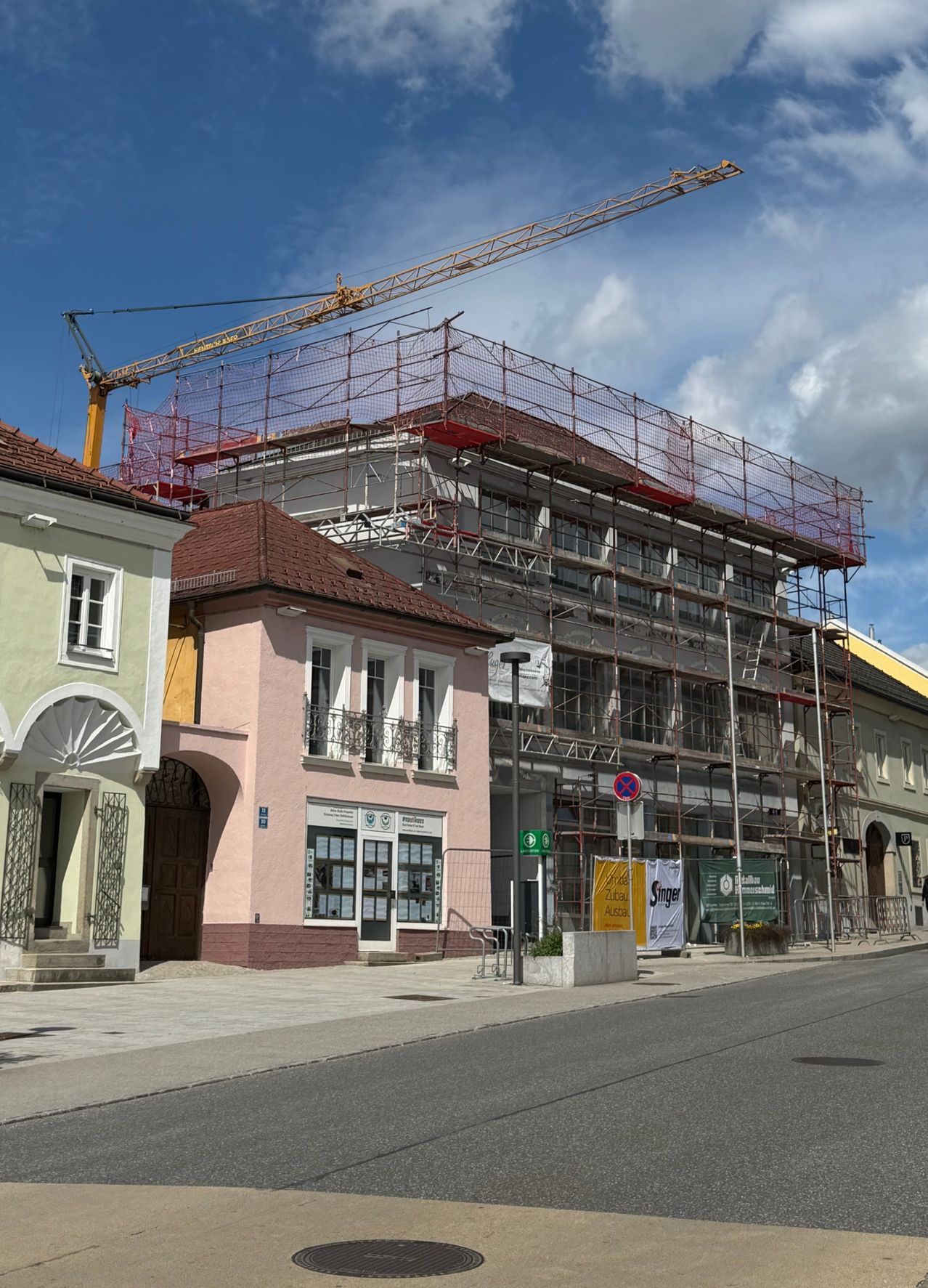 Eine Stadtstraße mit einem hohen Kran über einem im Bau befindlichen Gebäude. Ein kleineres Gebäude mit rosa Wänden und einem braunen Dach steht daneben.