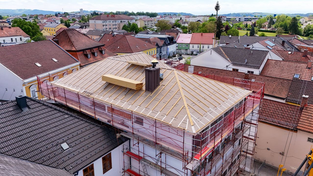 Ein Blick von oben auf eine Stadt zeigt ein Gebäude im Bau mit Gerüsten. Das Dach wird bearbeitet, mit Holzbrettern und einem Schornstein sichtbar. In der Nähe sind andere Gebäude mit verschiedenen Dachfarben zu sehen.