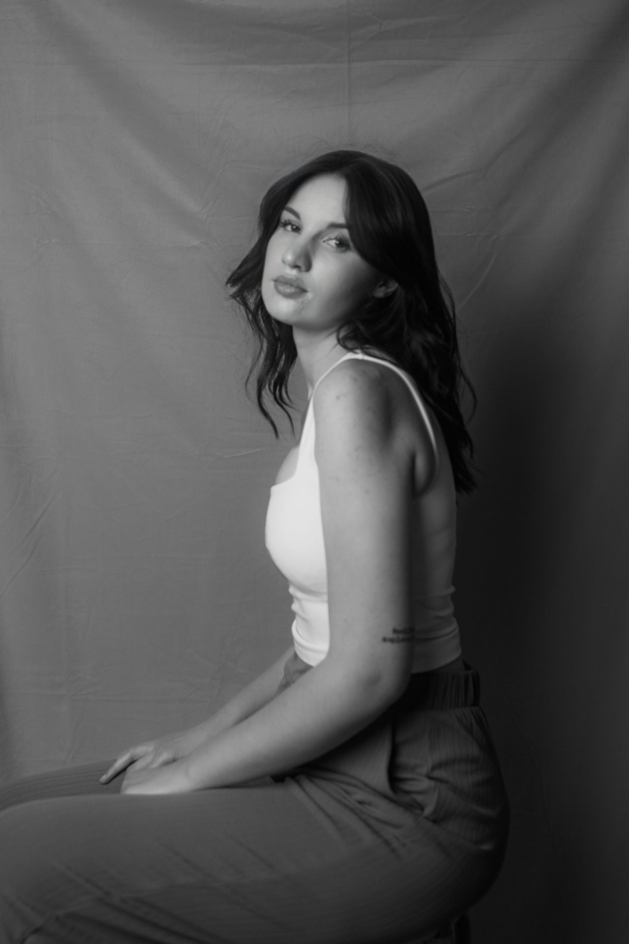 Ein schwarz-weißes Porträt einer Frau, die mit überkreuzten Beinen sitzt, ein weißes Tanktop und eine Hose trägt. Sie hat langes, dunkles Haar und eine Tätowierung am linken Arm. Sie schaut nach vorne mit leicht geöffnetem Mund.
