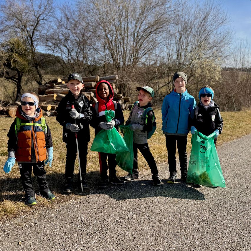 Eine Gruppe von Kindern, die Handschuhe tragen und Plastiktüten halten, posiert für ein Foto auf einer Straße. Sie stehen vor einigen kahlen Bäumen und einem Holzstapel.